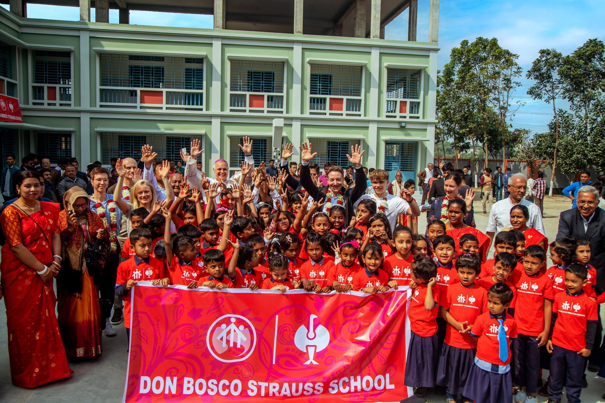 Group of children and adults in front of a school building with a Don Bosco Strauss School banner.